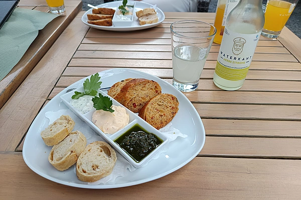 Dreiteiliger weißer Teller mit verschiedenen Brotscheiben und drei Dips auf einem Holztisch neben einem Glas Wasser und einer Flasche Getränk