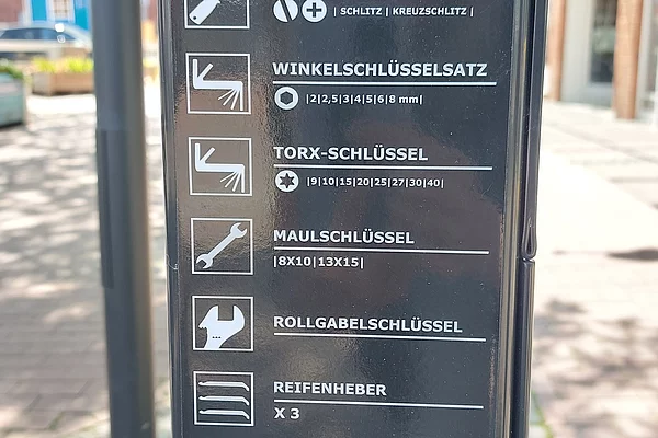 Schwarze Tafel mit weißen Piktogrammen und Texten zu Werkzeugen wie Winkelschlüssel, Torx-Schlüssel, Maulschlüssel, Rollgabelschlüssel, Reifenheber und Luftpumpe an einem Fahrradreparaturständer