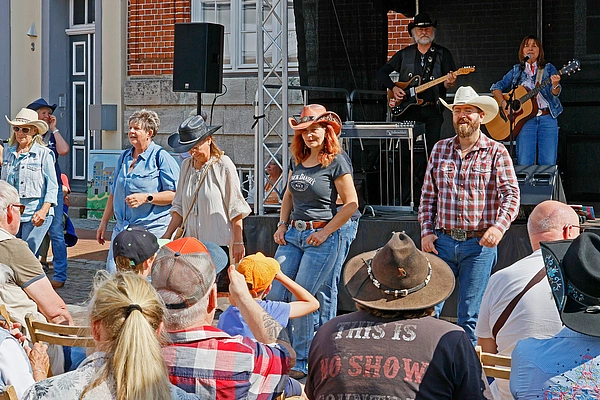 White Mountain Linedance Mehrere Menschen in Cowboyhüten tanzen vor einer Bühne, auf der zwei Musiker mit Gitarren spielen, Publikum sitzt im Vordergrund.