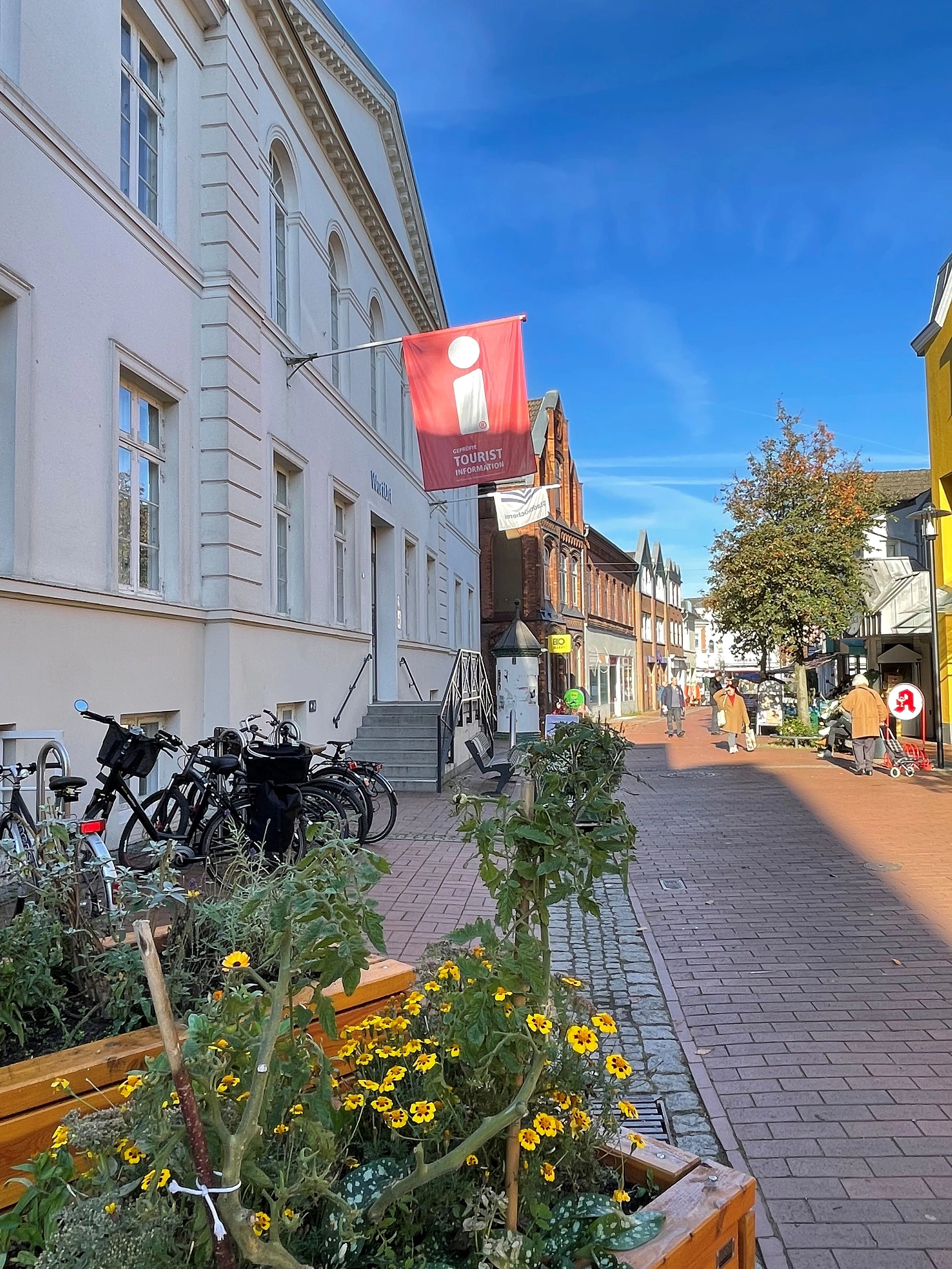 Straße in Bad Segeberg mit Fahrradständern, Blumenbeeten und einem roten Tourist-Informationsschild an einem weißen Gebäude