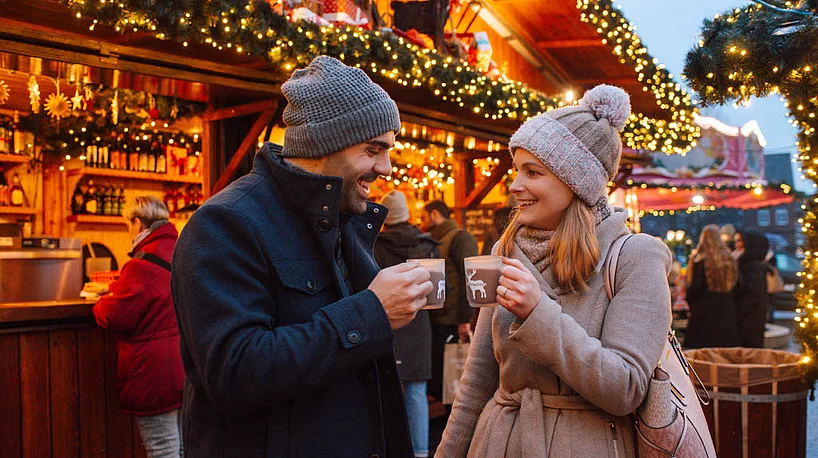 Zwei Personen in Winterkleidung mit Mützen halten Tassen vor einem festlich beleuchteten Holzstand auf dem Weihnachtsmarkt Bad Segeberg