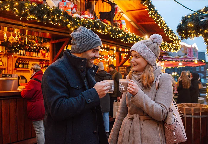 Zwei Personen in Winterkleidung mit Mützen halten Tassen vor einem festlich beleuchteten Holzstand auf dem Weihnachtsmarkt Bad Segeberg