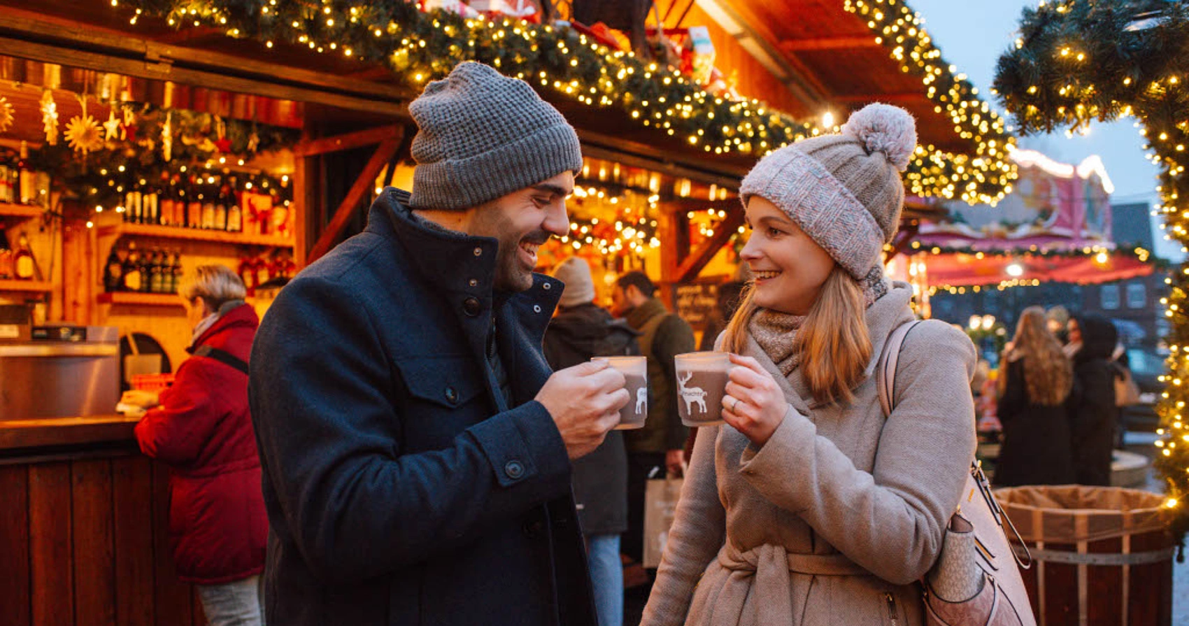 Zwei Personen in Winterkleidung mit Mützen halten Tassen vor einem festlich beleuchteten Holzstand auf dem Weihnachtsmarkt Bad Segeberg