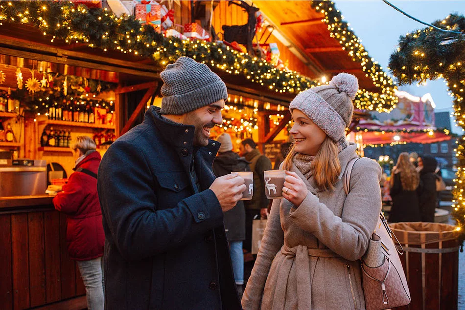 Zwei Personen in Winterkleidung mit Mützen halten Tassen vor einem festlich beleuchteten Holzstand auf dem Weihnachtsmarkt Bad Segeberg