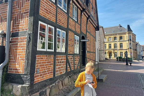 Mädchen mit gelber Jacke steht vor einem Fachwerkhaus an einer gepflasterten Straße in einer historischen Altstadt
