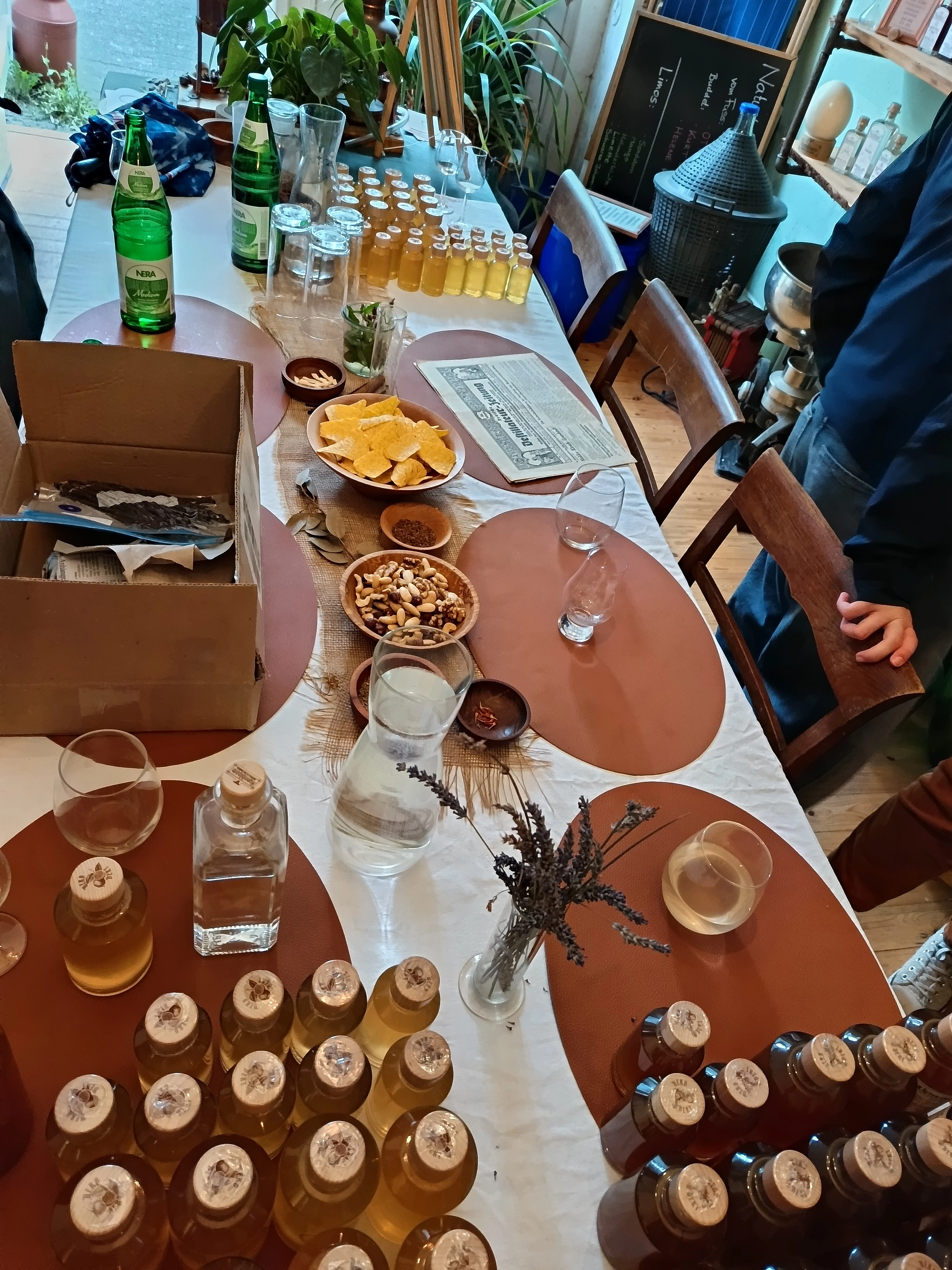 Tisch mit mehreren Flaschen, Gläsern, Schalen mit Snacks und einer Zeitung, daneben Stühle und eine stehende Person mit dunkler Kleidung