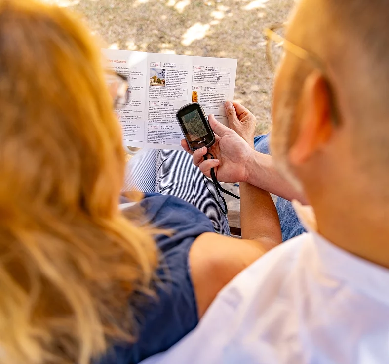 Zwei Personen von hinten sitzend, eine hält ein GPS-Gerät und eine Faltkarte mit Geocaching-Informationen