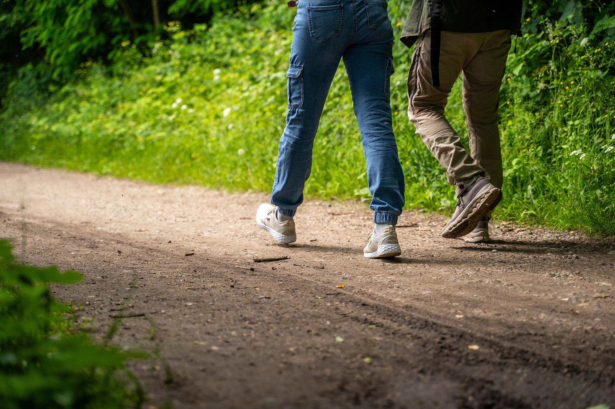 Zwei Personen gehen auf einem Waldweg, sichtbar sind nur ihre Beine und Füße in Jeans und Freizeitschuhen