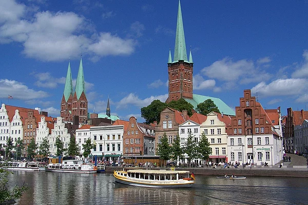 Blick auf die historische Altstadt von Lübeck mit mehreren Kirchen und Giebelhäusern am Wasser, davor ein Ausflugsboot auf dem Fluss