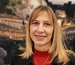 Frau mit schulterlangem blonden Haar trägt roten Rollkragenpullover und schwarze Jacke vor unscharfem Hintergrund 