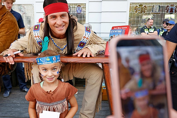 Ein Mann in Indianerkostüm mit rotem Stirnband lehnt an einem Tisch und lächelt in die Kamera, vor ihm steht ein Mädchen mit blauem Kopfschmuck und braunem Kleid, im Vordergrund ist ein Smartphone zu sehen, das die Szene filmt.