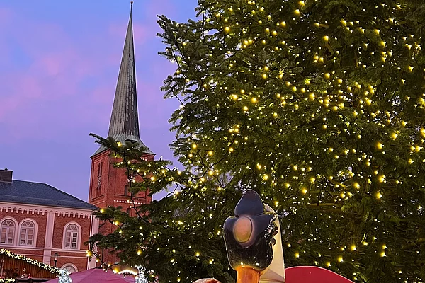 Weihnachtlich geschmückter großer Tannenbaum mit gelben Lichtern vor rotem Backsteingebäude und Kirchturmspitze bei Dämmerung, davor eine weiße Schwan-Skulptur mit rotem Sitz.