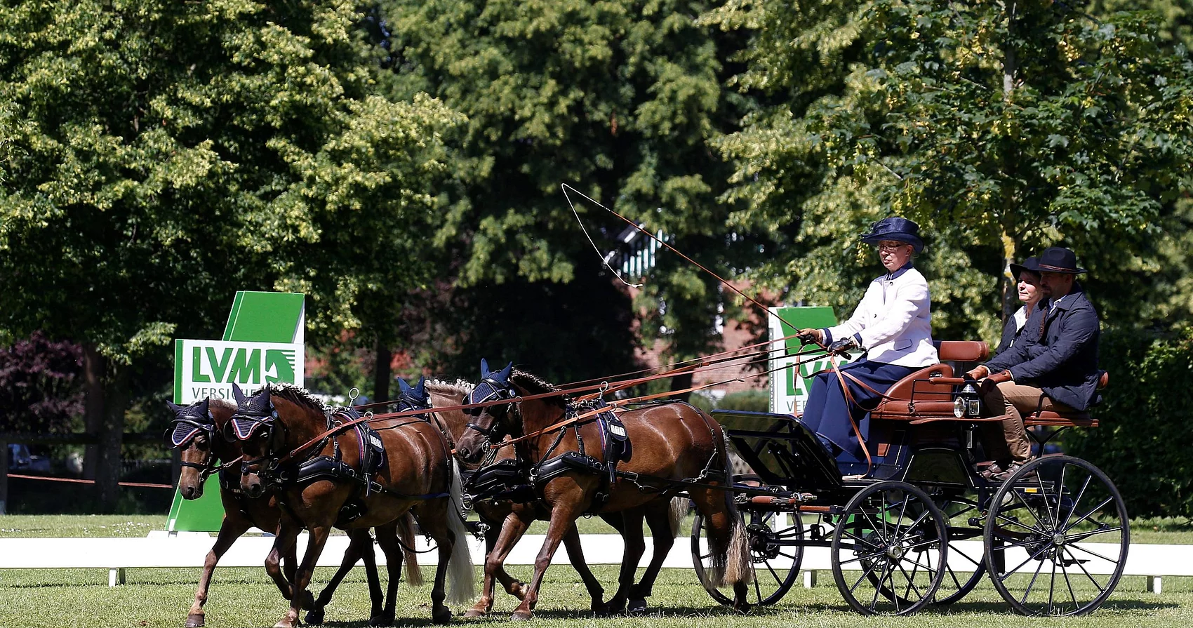 Vier Pferde ziehen eine Kutsche mit drei Personen auf einer Wiese vor Bäumen und Werbetafeln mit dem Schriftzug 'LVM Versicherung'