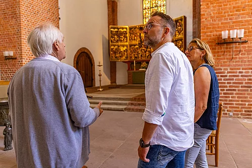 Drei Personen bei einer Stadtführung in einer Kirche mit rotem Backsteinmauerwerk und goldenem Altar im Hintergrund
