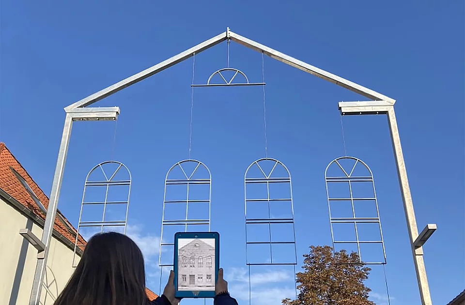 Person mit langen dunklen Haaren hält ein Tablet vor eine Stahlrahmenkonstruktion in Form von Fenstern und einem Giebel, im Hintergrund Häuser und blauer Himmel