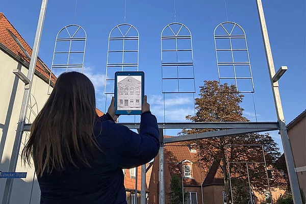 Person mit langen dunklen Haaren hält ein Tablet vor eine Stahlrahmenkonstruktion in Form von Fenstern und einem Giebel, im Hintergrund Häuser und blauer Himmel