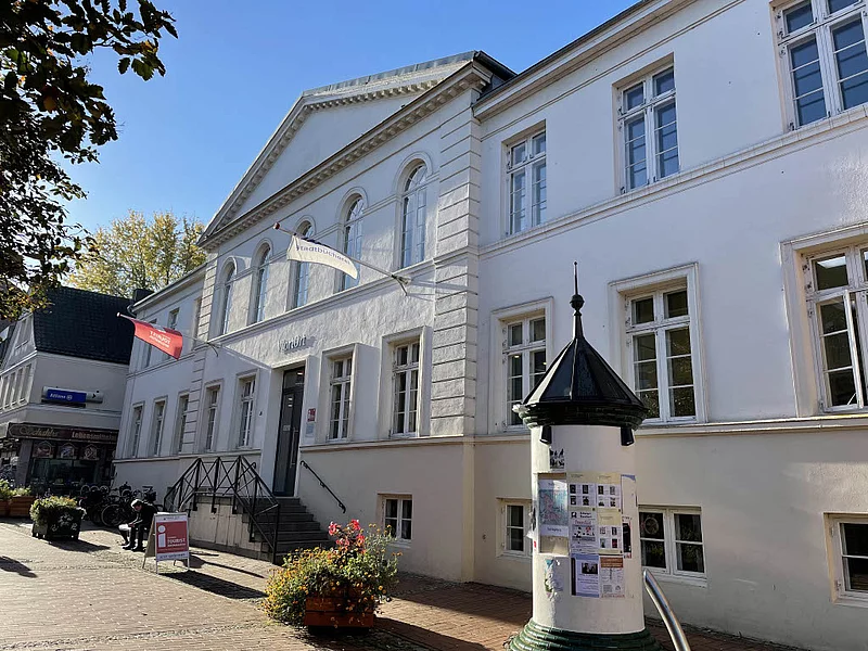 Weißes klassizistisches Gebäude mit Treppe und zwei wehenden Fahnen vor blauem Himmel