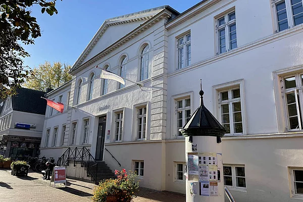 Weißes klassizistisches Gebäude mit Treppe und zwei wehenden Fahnen vor blauem Himmel