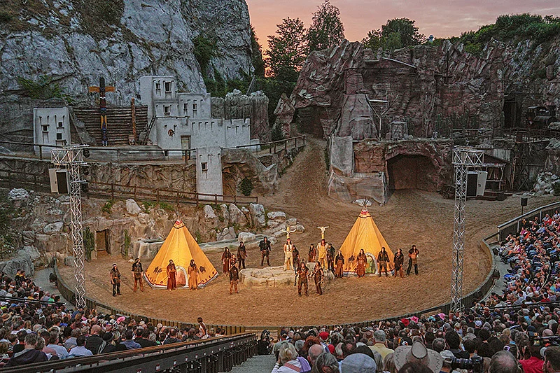 Freilichtbühne der Karl-May-Spiele Bad Segeberg mit Schauspielern in Indianer- und Westernkostümen vor zwei beleuchteten Tipis und Felsenbühne, umgeben von Zuschauerrängen mit Publikum