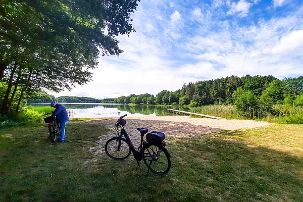 Fahrräder auf einer Wiese am Ufer eines Sees mit Wald und blauem Himmel im Hintergrund