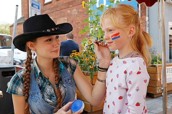 Junge Frau mit Cowboyhut malt einem Mädchen mit roten und blauen Streifen auf der Wange das Gesicht an