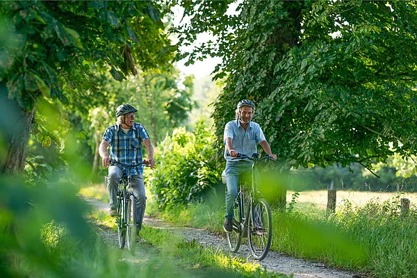 Zwei Männer mit Fahrradhelmen fahren auf einem schmalen Waldweg umgeben von Bäumen und Grün