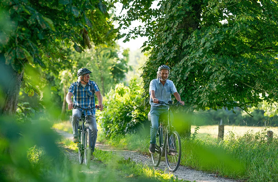 Zwei Männer mit Fahrradhelmen fahren auf einem schmalen Waldweg umgeben von Bäumen und Grün