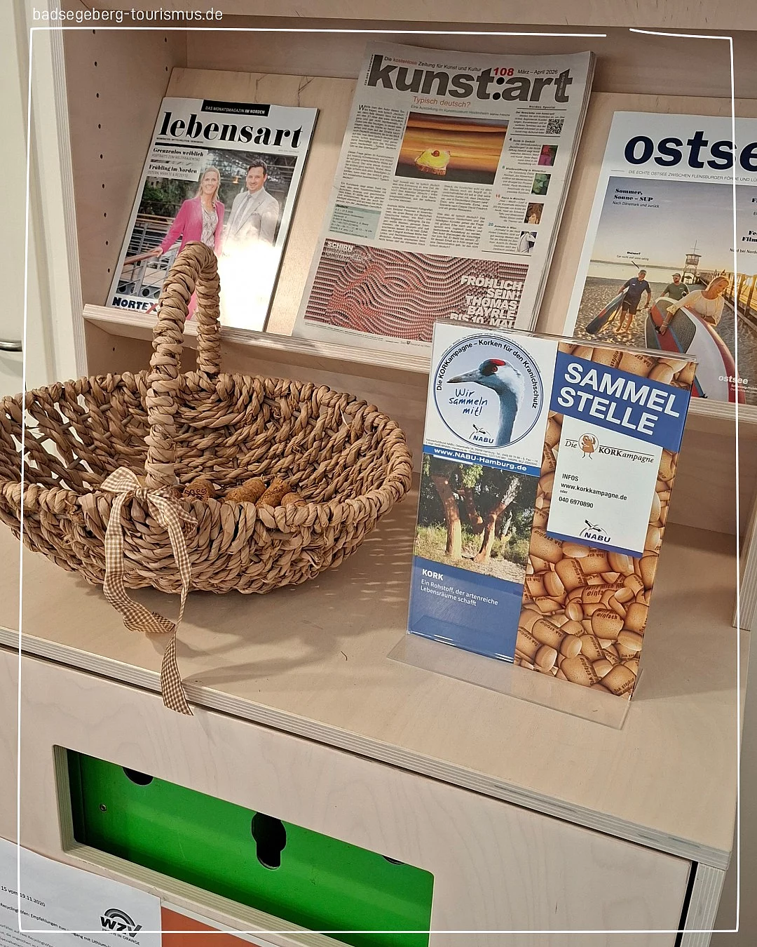 Geflochtener Korb mit Griff und Schleife steht auf einem hellen Möbelstück vor mehreren aufgestellten Zeitungen und einem Informationsaufsteller mit Korkenmotiv