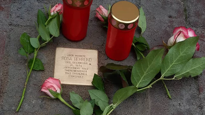 Stolperstein mit eingraviertem Namen und Daten, umgeben von drei Rosen und zwei roten Gedenkkerzen mit goldfarbenen Deckeln auf Pflastersteinen