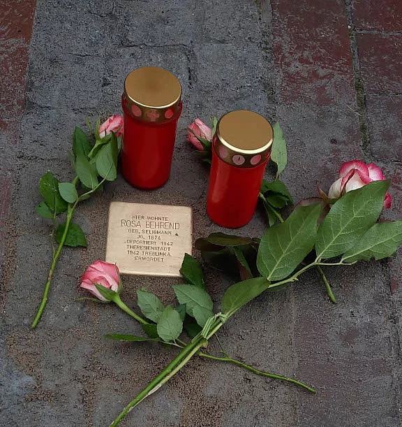 Stolperstein mit eingraviertem Namen und Daten, umgeben von drei Rosen und zwei roten Gedenkkerzen mit goldfarbenen Deckeln auf Pflastersteinen