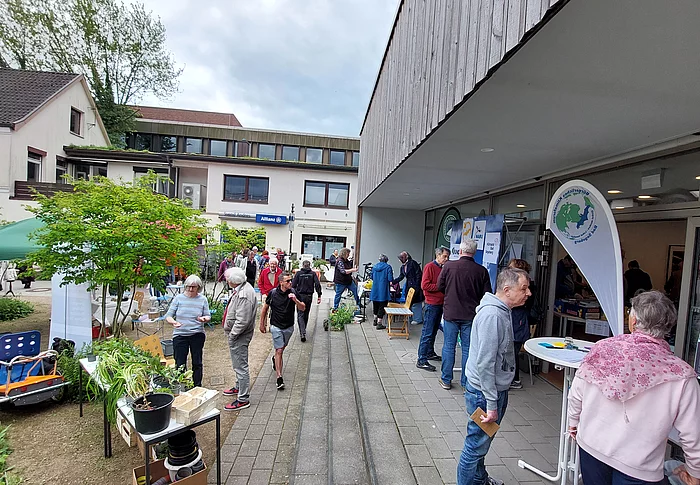 Menschen auf einem Gehweg vor einem Gebäude mit Informationsständen und einem Banner mit grünem Logo
