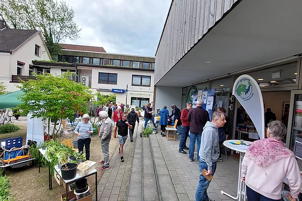 Menschen auf einem Gehweg vor einem Gebäude mit Informationsständen und einem Banner mit grünem Logo