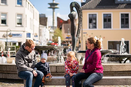 Familie mit zwei Kindern sitzt auf einer Steinbank vor dem Marktbrunnen Bad Segeberg mit Wasserspielen und einer Skulptur im Hintergrund