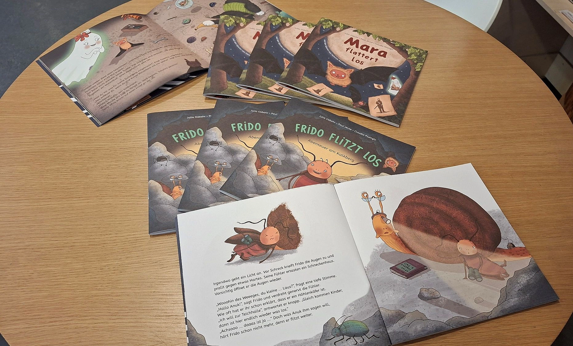 Auf einem runden Holztisch liegen mehrere aufgeschlagene und geschlossene Kinderbücher mit Illustrationen von Tieren und Texten.