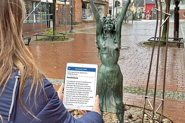 Bronzefigur einer Frau mit erhobenen Armen, die mit einer Flüssigkeit übergossen wird, auf einem runden Pflastersteinboden in einem städtischen Umfeld mit Backsteingebäuden und Bäumen im Hintergrund