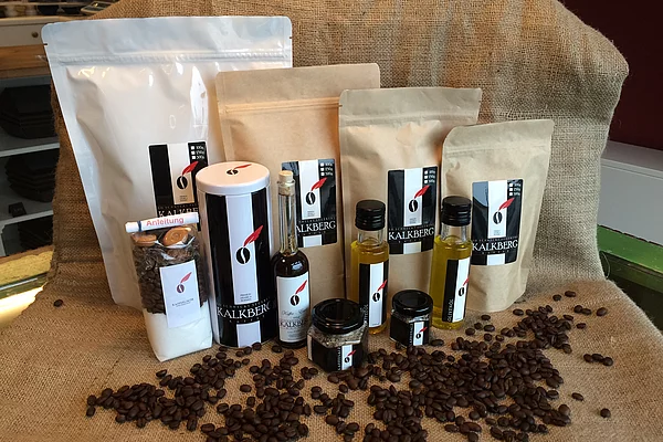 Verschiedene Verpackungen und Flaschen mit KALKBERG-Logo, umgeben von Kaffeebohnen auf einem Stoffhintergrund