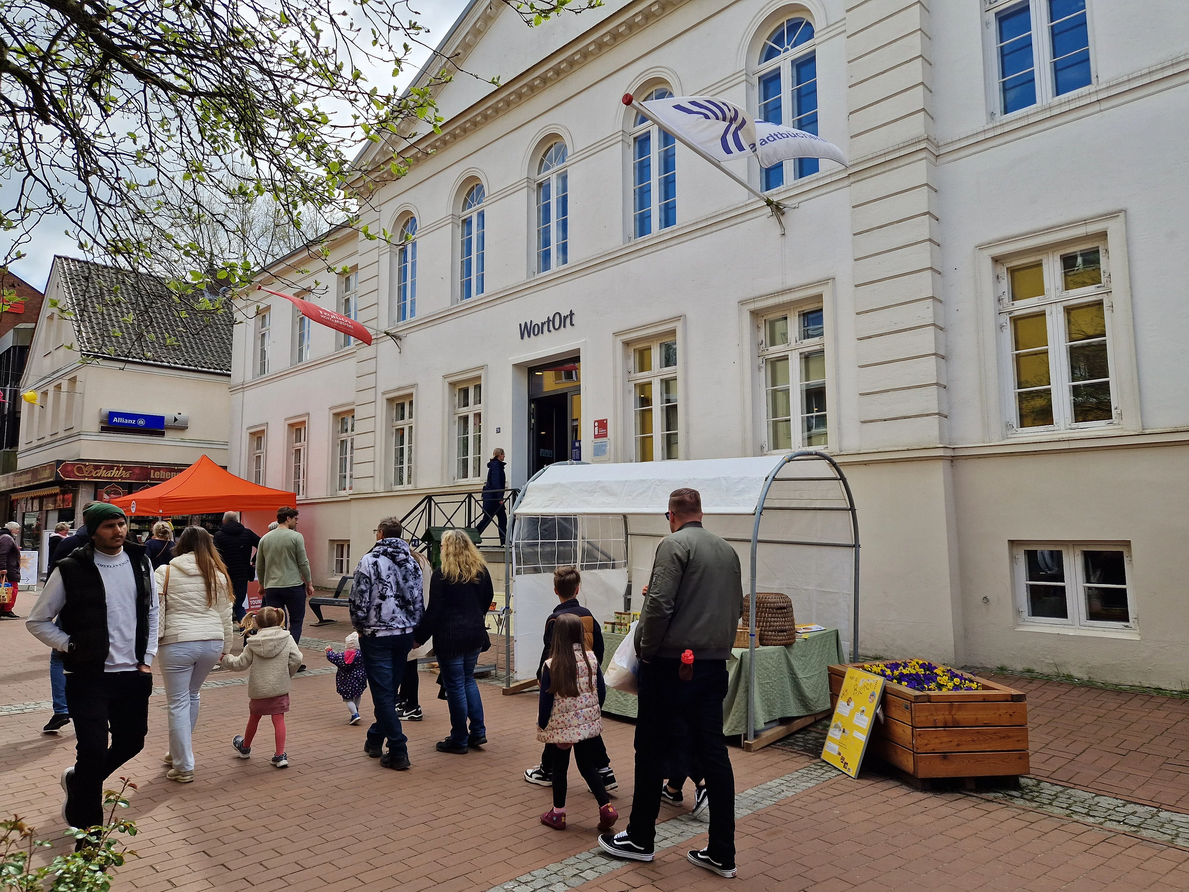 Menschen gehen auf einem gepflasterten Platz an einem weißen Gebäude mit der Aufschrift 'WortOrt' vorbei, davor ein Stand mit einem weißen Pavillon und Blumenkästen