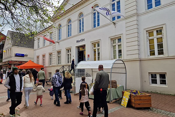 Menschen gehen auf einem gepflasterten Platz an einem weißen Gebäude mit der Aufschrift 'WortOrt' vorbei, davor ein Stand mit einem weißen Pavillon und Blumenkästen
