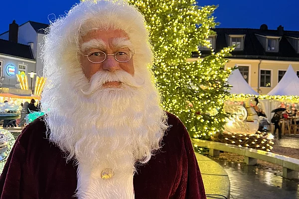 Weihnachtsmann mit rotem Samtmantel und weißem Haar und Bart vor einem großen, mit Lichtern geschmückten Weihnachtsbaum auf einem festlich beleuchteten Marktplatz in Bad Segeberg