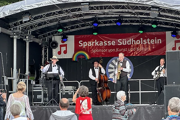 Vier Musiker auf einer Bühne, im Hintergrund ein rotes Banner mit der Aufschrift 'Sparkasse Südholstein Sponsor von Kunst und Kultur', davor tanzen mehrere Zuschauer.