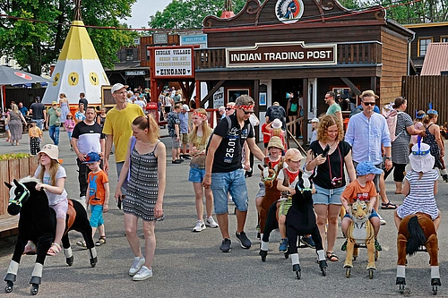 Menschen verschiedener Altersgruppen gehen und reiten auf Spielpferden im Indian Village von den Karl-May-Spielen mit einem Gebäude namens 'Indian Trading Post' und einem Tipi im Hintergrund