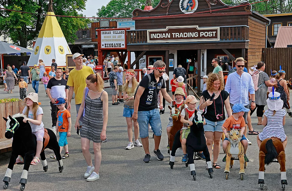 Menschen verschiedener Altersgruppen gehen und reiten auf Spielpferden im Indian Village von den Karl-May-Spielen mit einem Gebäude namens 'Indian Trading Post' und einem Tipi im Hintergrund
