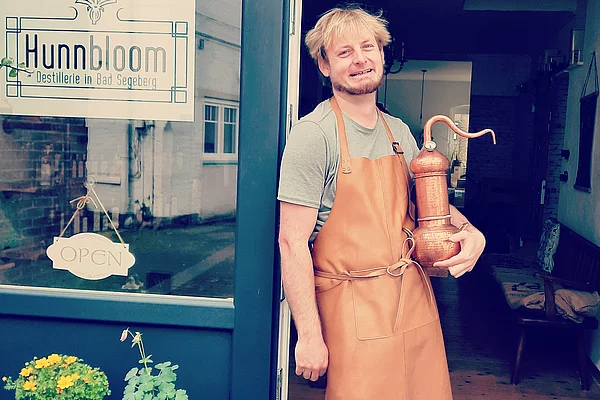 Person mit brauner Lederschürze steht in einem Türrahmen und hält eine kupferfarbene Destillationsapparatur