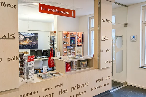 Innenraum einer Tourist-Information mit Theke, Informationsmaterial und Wänden, an denen Wörter in verschiedenen Sprachen stehen