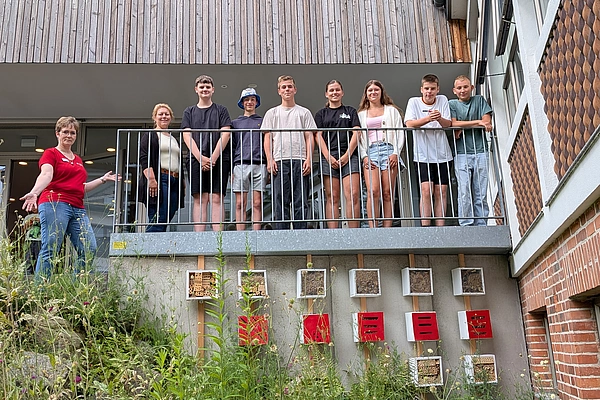 Gruppe von Jugendlichen und einer Frau steht auf einem Balkon eines Gebäudes mit Holzverkleidung und begrüßt mit ausgestrecktem Arm