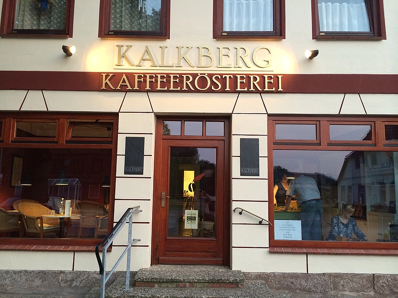 Fassade eines Gebäudes mit dem Schriftzug 'KALKBERG KAFFEERÖSTEREI' über einem zentralen Eingang mit Glastür und großen Fenstern, durch die Menschen im Inneren sichtbar sind