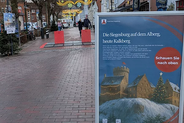 Fußgängerzone mit Pflastersteinen, im Vordergrund ein Informationsschild mit Bild und Text zur Siegesburg auf dem Kalkberg, im Hintergrund Häuser, Bäume ohne Blätter und Menschen auf der Straße
