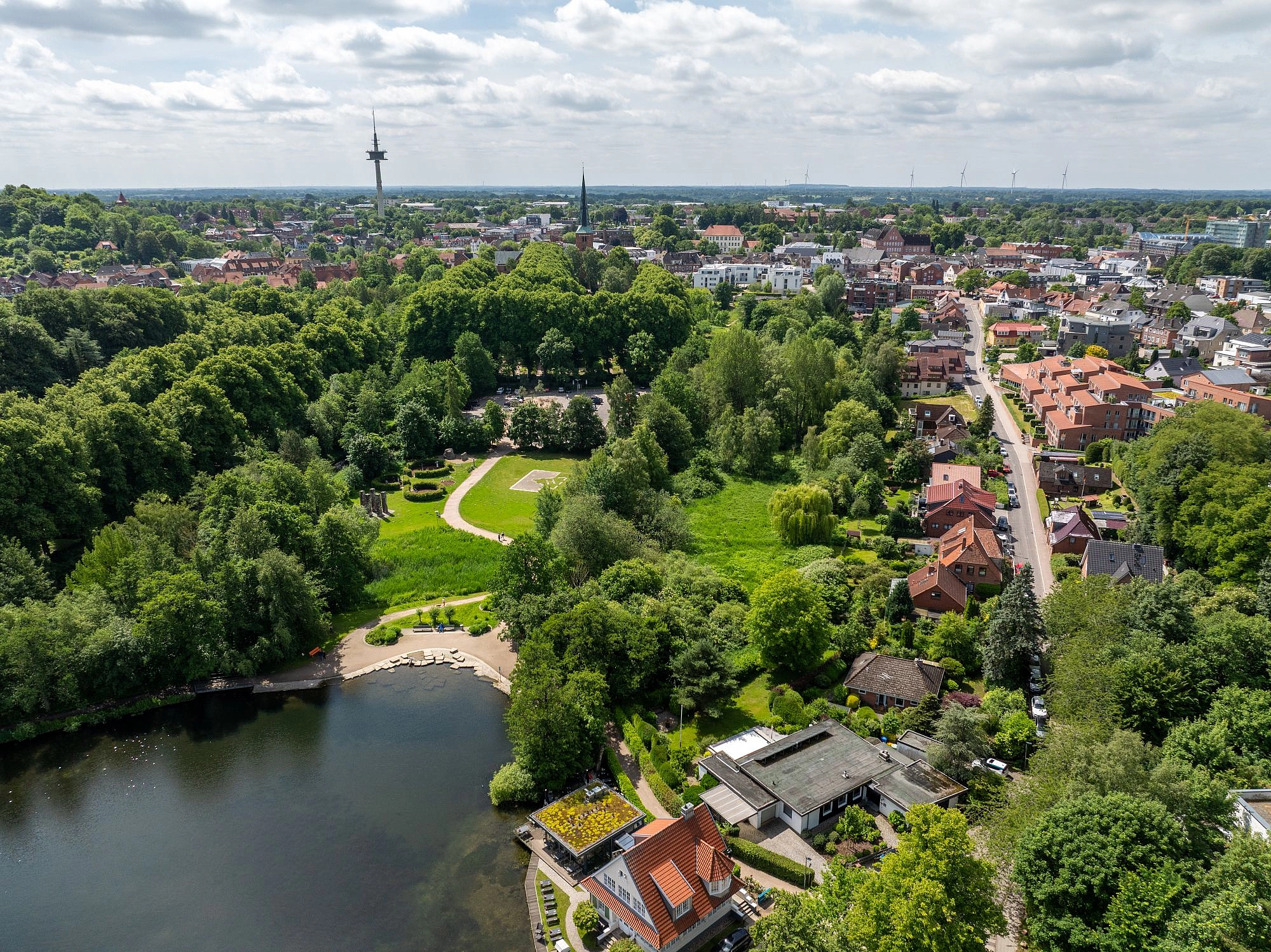 Luftaufnahme eines Parks mit See, umgeben von dichtem Grün und angrenzenden Wohnhäusern in einer Stadt