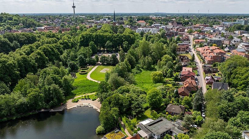 Luftaufnahme eines Parks mit See, umgeben von dichtem Grün und angrenzenden Wohnhäusern in einer Stadt