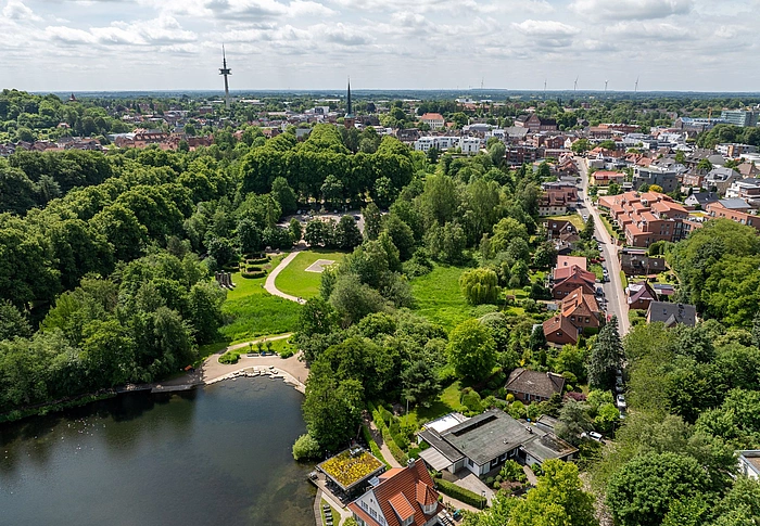 Luftaufnahme eines Parks mit See, umgeben von dichtem Grün und angrenzenden Wohnhäusern in einer Stadt